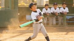 野球のユニフォームを着た少年が満面の笑みでバットを振っており土煙が舞っている背景には他の少年たちが笑顔で見守っている